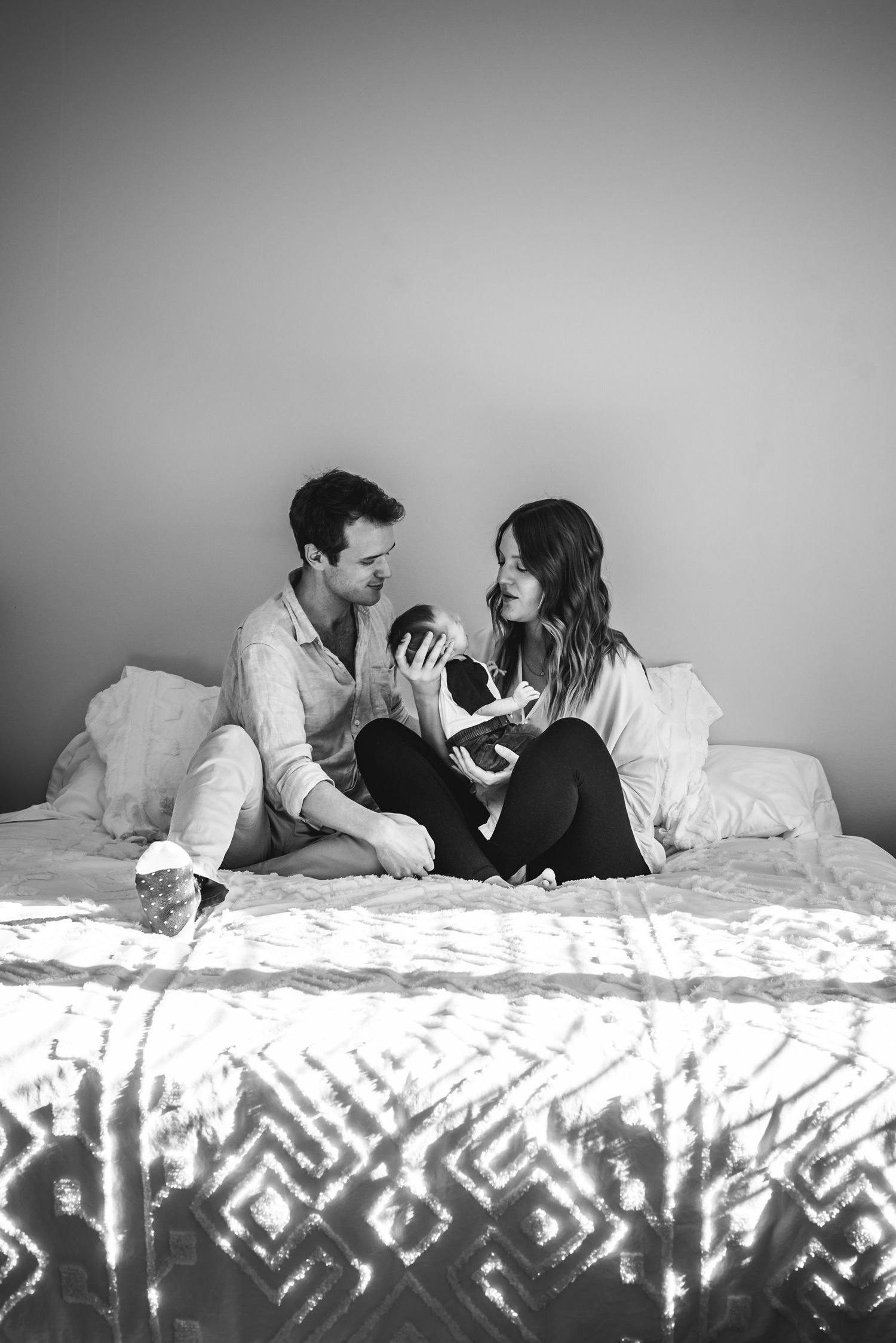 BW image of a man and woman sitting on a bed holding their newborn son.