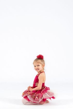 Young performer in red tutu dress sits in ballet pose on white background.