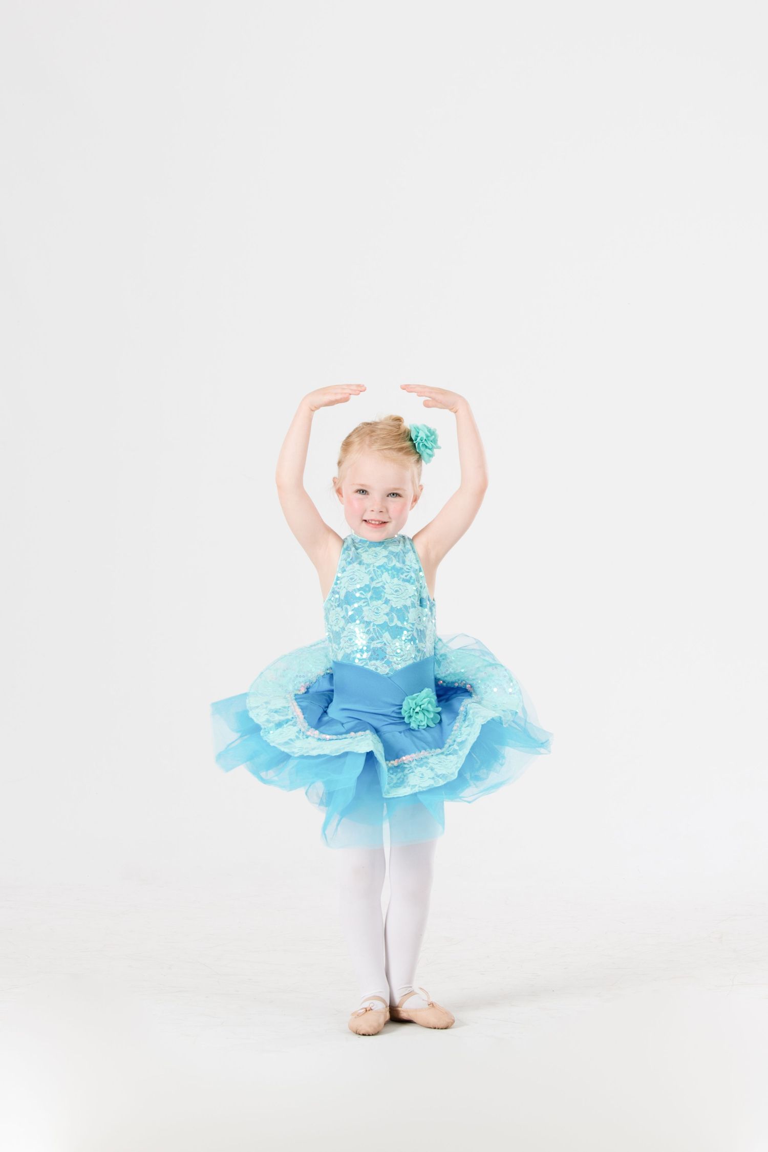 Young dancer in light blue tutu with ruffles strikes graceful ballet pose against white background.