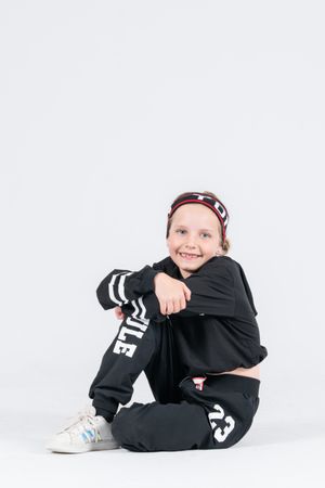 A child in black athletic wear and a patterned headband sits on the floor in a relaxed pose against a white backdrop.