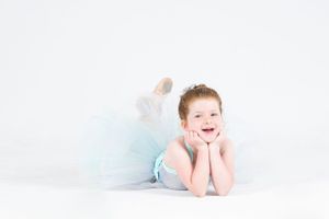 Young dancer in a pale blue tutu rests on the floor against a white background with a cheerful expression.