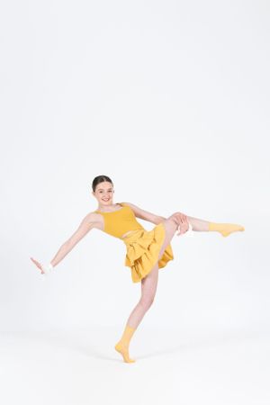 A dancer in a yellow dress and socks strikes a dynamic pose with one leg extended against a white background.