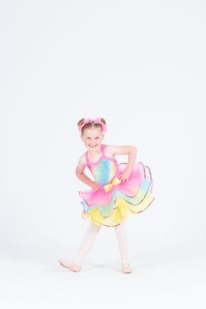 Young dancer poses playfully in colorful tutu dress with blue and pink accents against white backdrop.