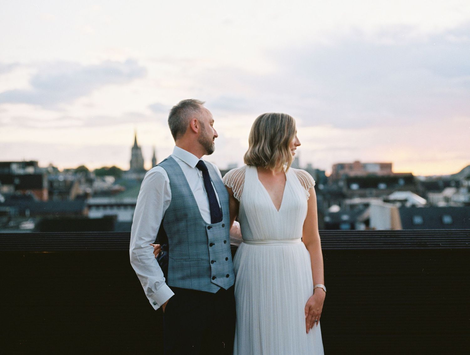 A couple in wedding attire admires a city skyline view from a rooftop during sunset.