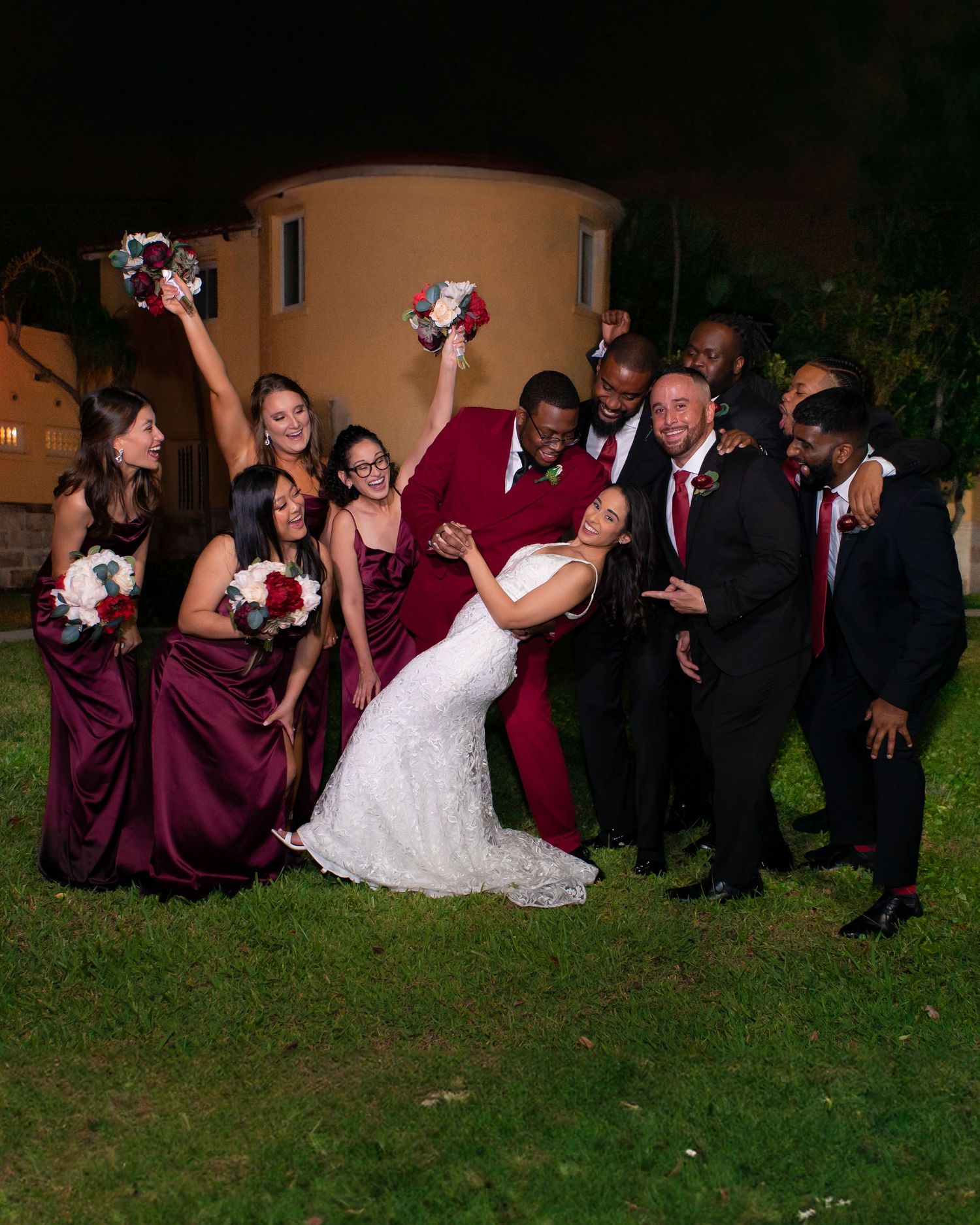 Wedding party celebrating outdoors at night in burgundy and white formal attire.