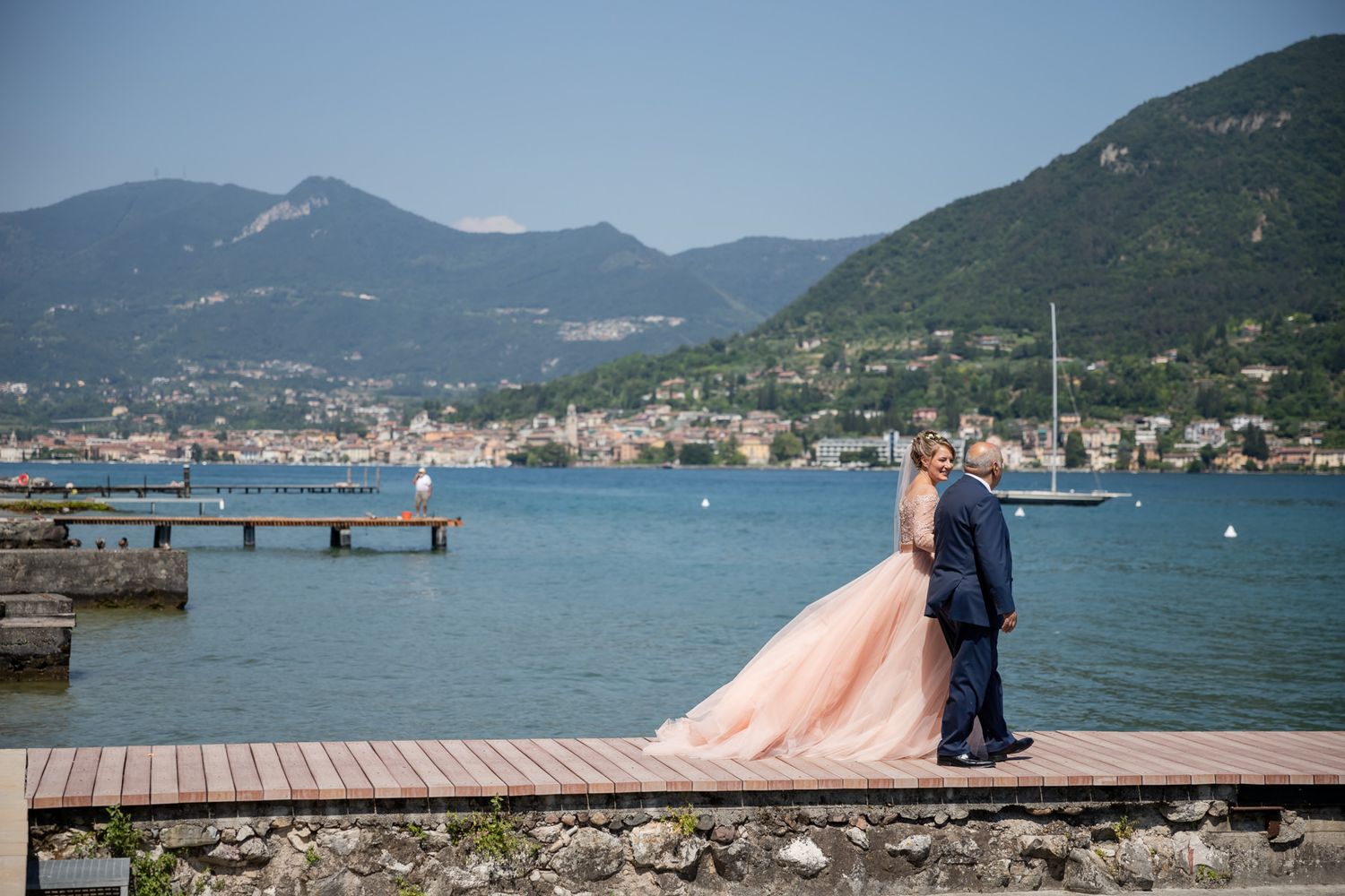 Scenic lakeside pier with mountains and town in background on a sunny day.