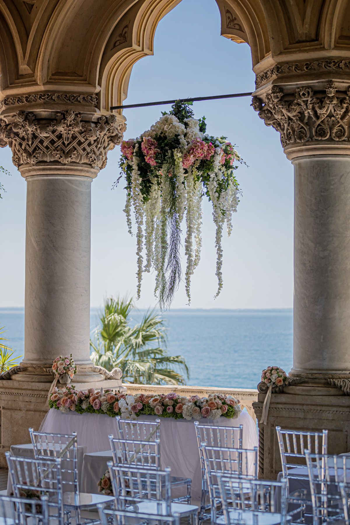 Oceanfront wedding ceremony setup with white chairs and floral decorations under ornate arches.