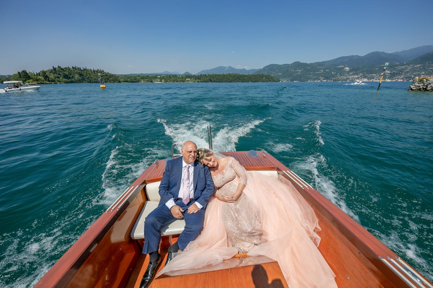 Couple riding in classic wooden boat across blue lake waters with mountains in background.