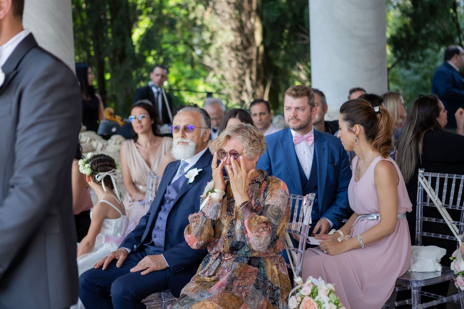 Wedding guests seated in white chairs during an outdoor ceremony under natural greenery.
