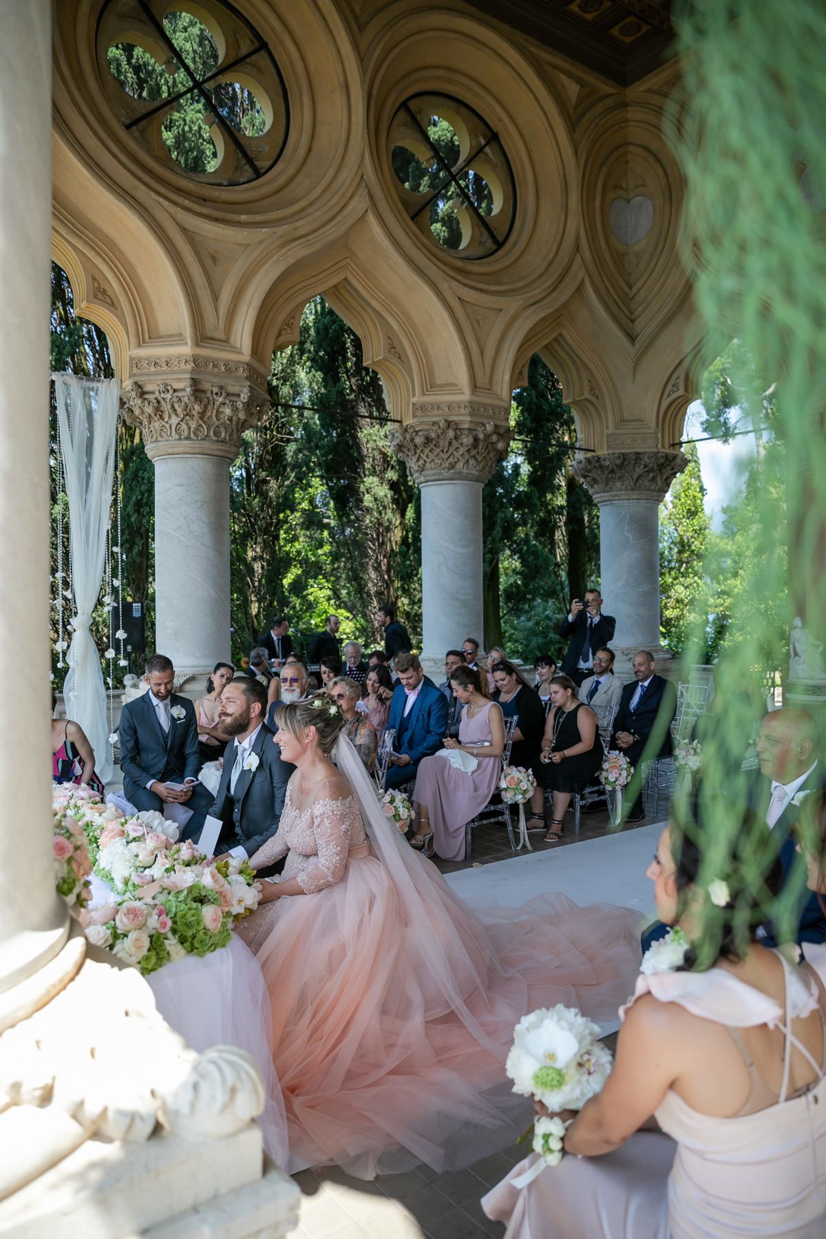 Wedding ceremony taking place under ornate stone columns with guests seated in an elegant outdoor venue.