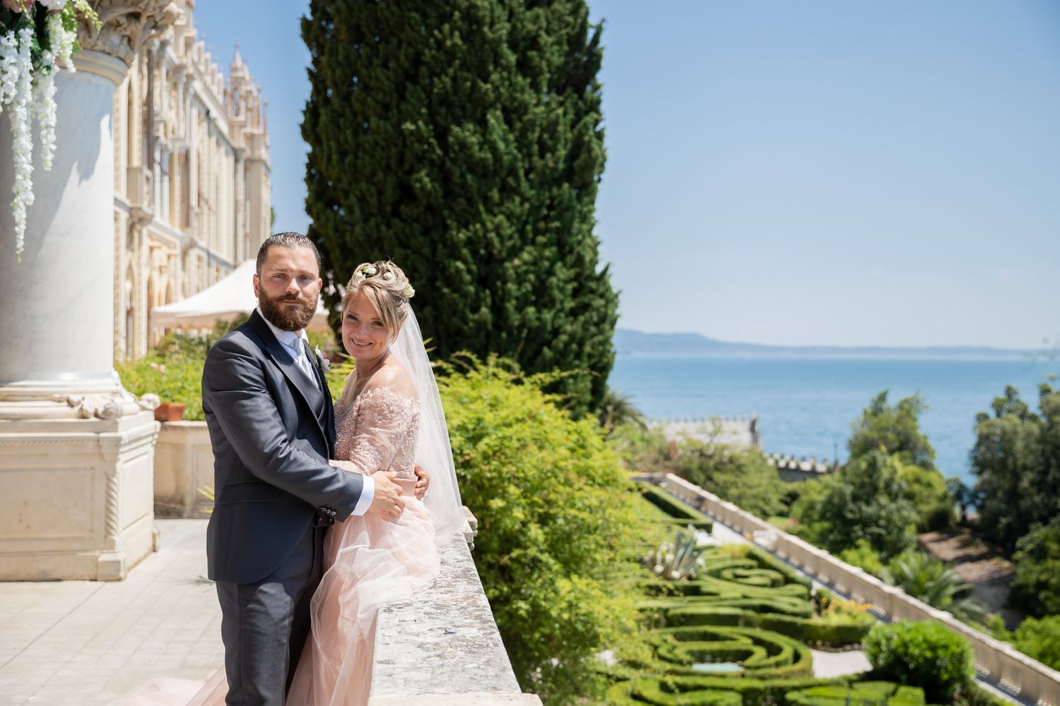 Elegant wedding couple embraces on a scenic terrace overlooking water and gardens.