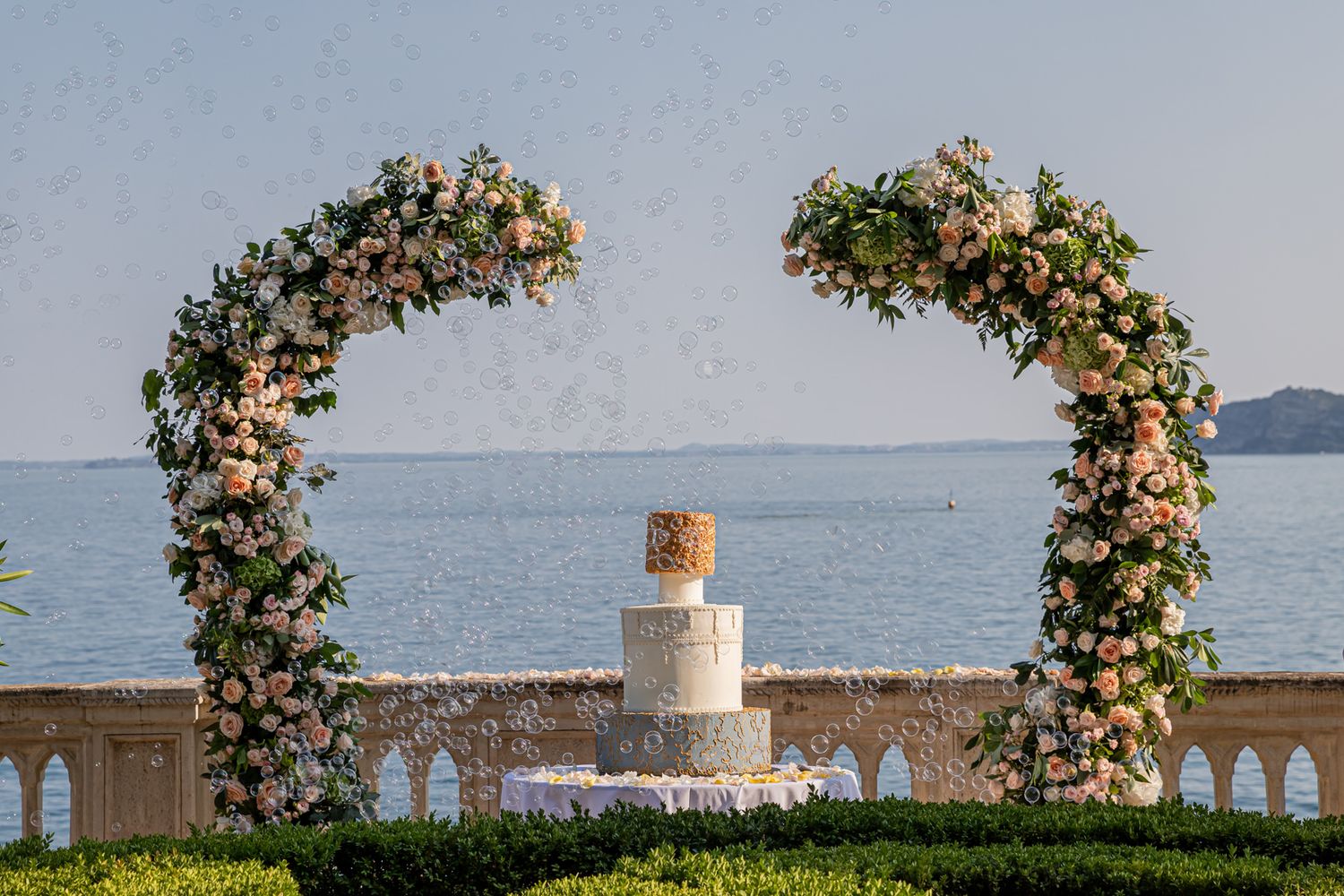 Elegant wedding cake display with floral arch decorations overlooking a scenic waterfront view.