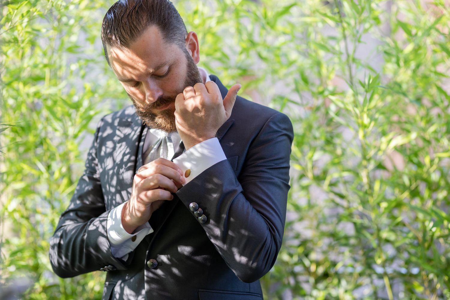 Man in suit adjusting tie outdoors with natural greenery background.
