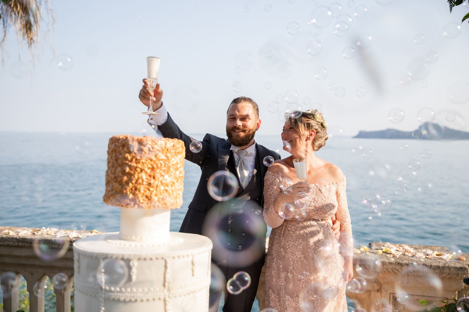 Wedding celebration with soap bubbles floating around an elegant tiered cake by the sea.