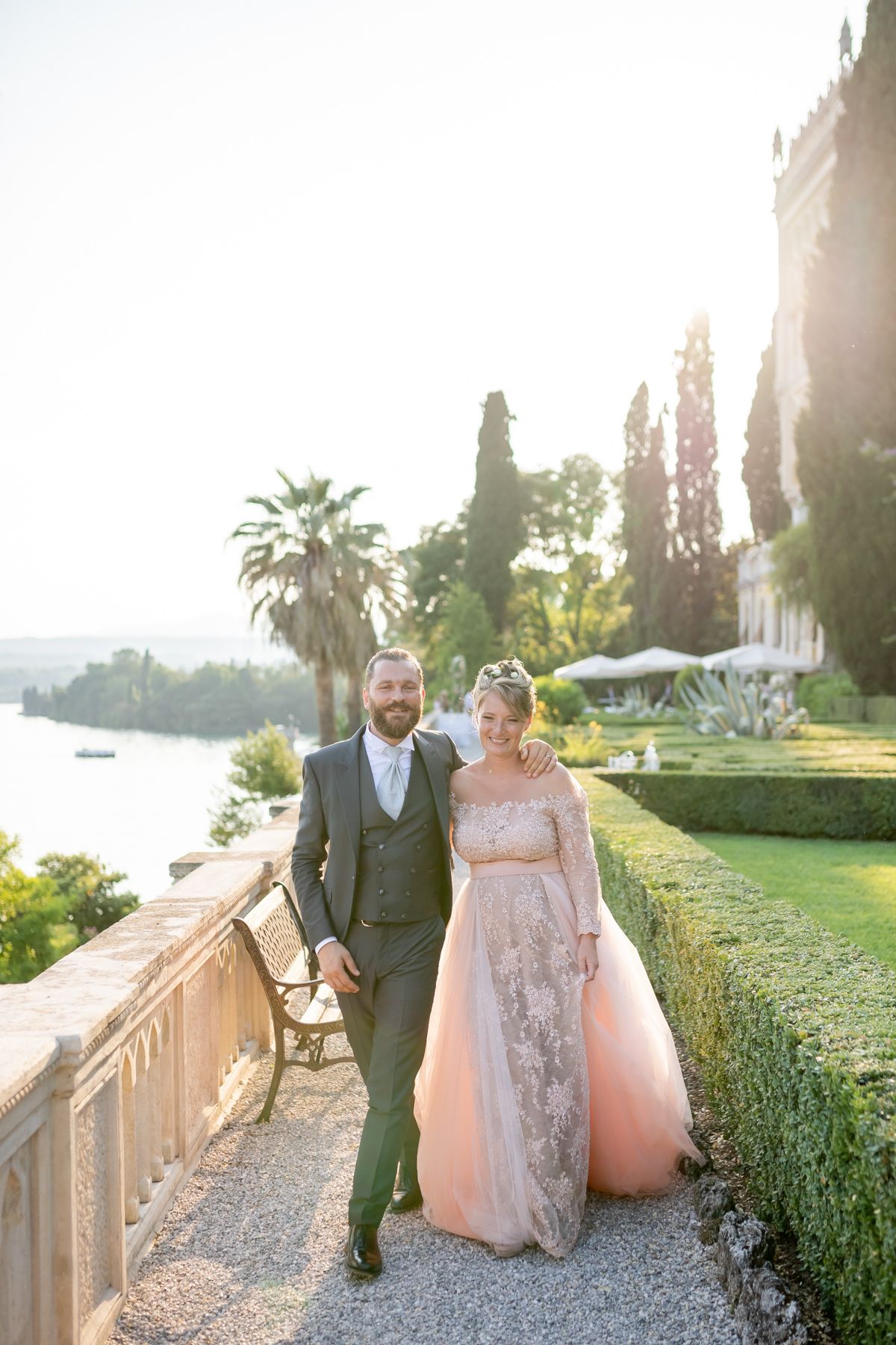 A couple walks along a stone balustrade at sunset in a Mediterranean garden setting with palm trees.