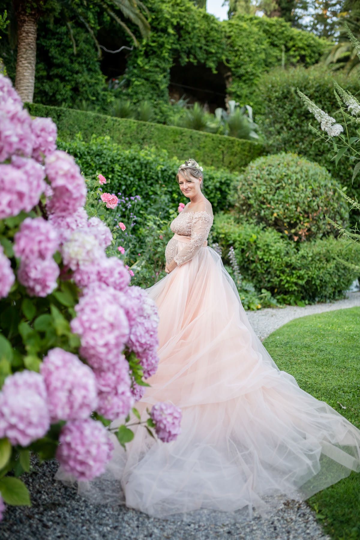 Blush pink wedding gown flowing dramatically among pink flowering bushes in a lush garden.