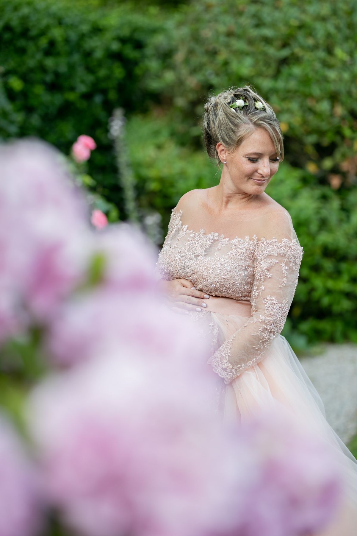 Close-up portrait through pink flowers showing delicate lace details of a blush wedding gown.
