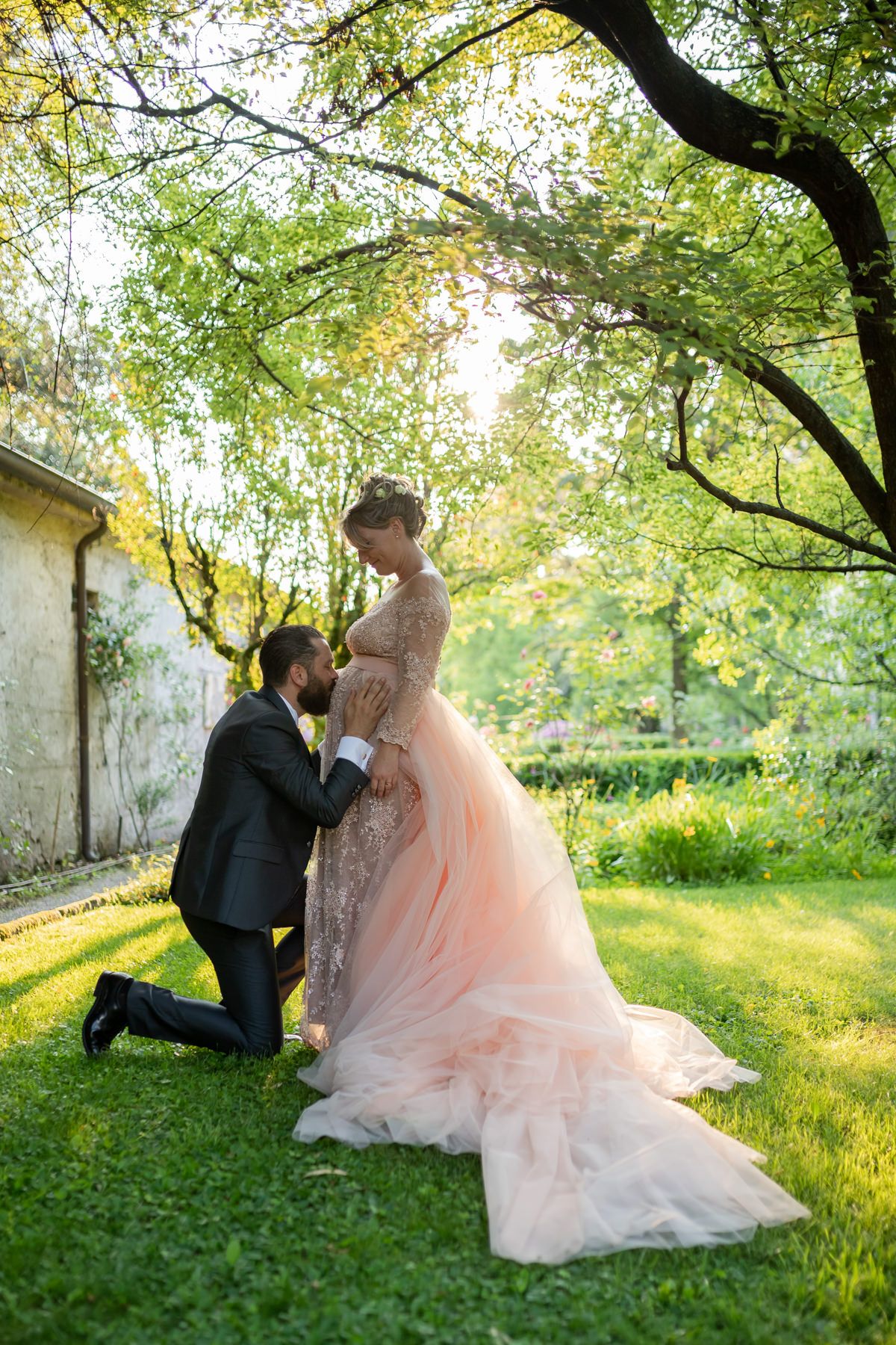 Couple shares tender moments on grassy lawn under arching tree branches at sunset.