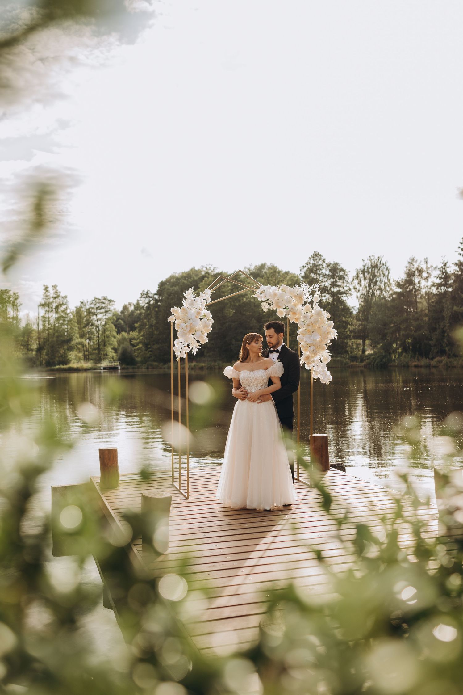A romantic lakeside wedding ceremony setup with white floral arch and wooden dock surrounded by nature.