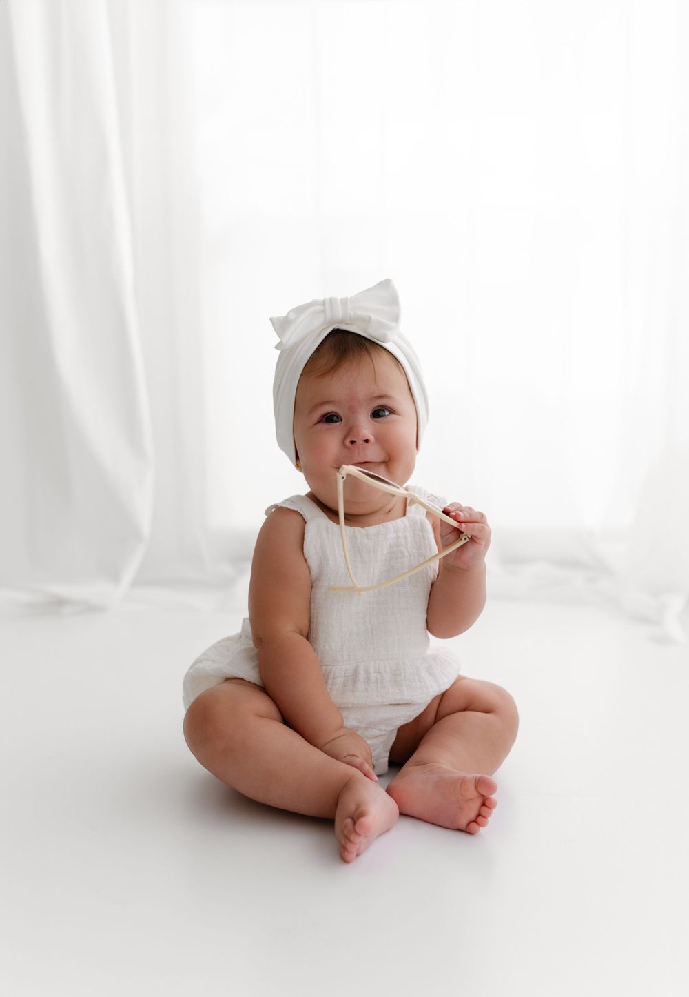 Baby in white outfit with bow headband sits and plays during minimalist studio session.