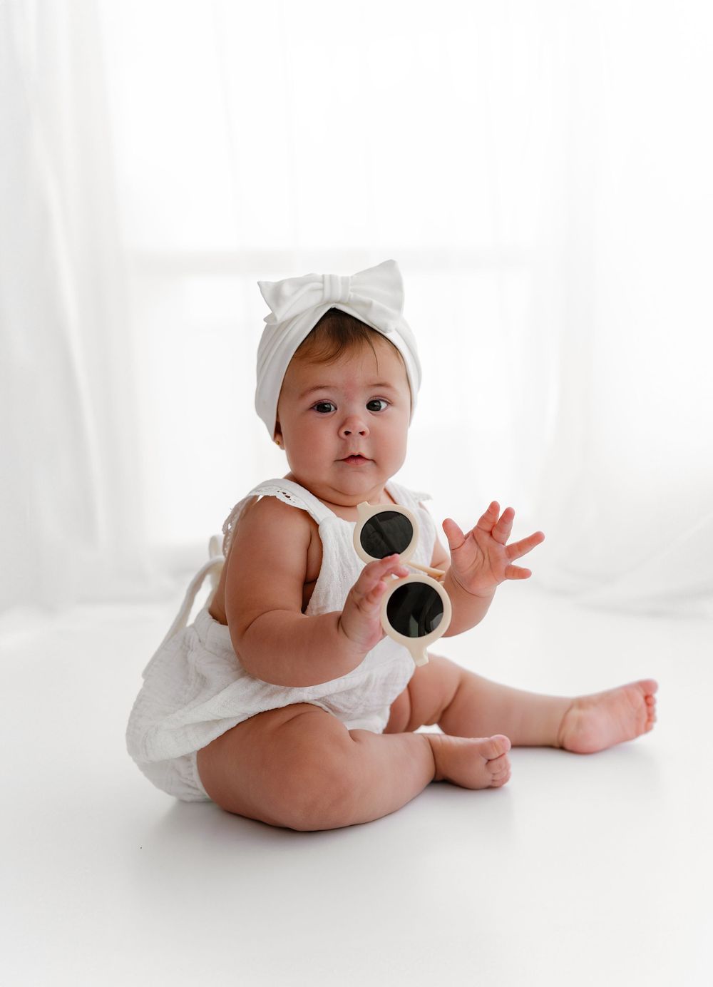 Baby wearing white romper and bow headband sitting with sunglasses against white background.