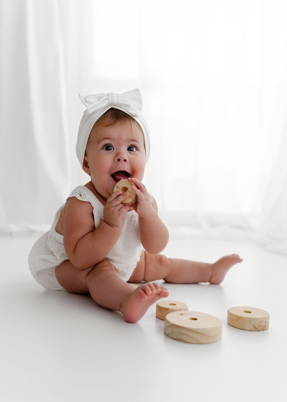 Baby in white outfit playing with wooden toys while sitting against white backdrop.