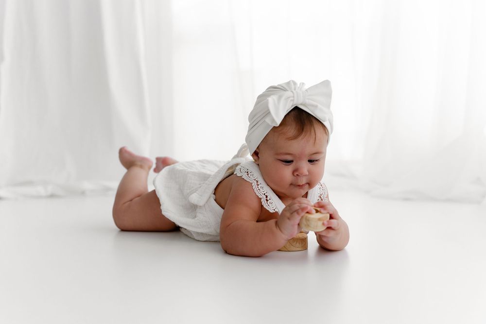 Baby in white dress lying on stomach playing with wooden toys on white floor.