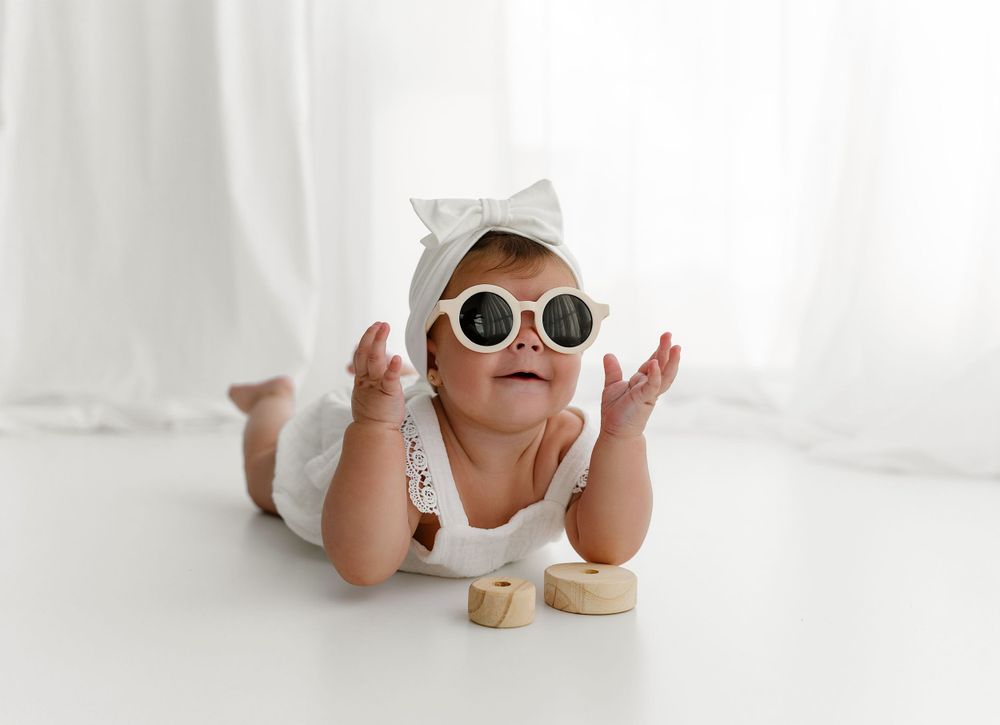 Baby wearing sunglasses and white outfit lies on stomach during playful studio photoshoot.