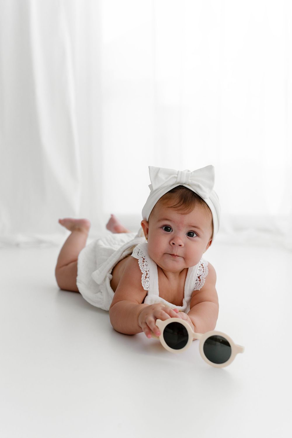 Baby in white outfit with bow headband lying on stomach while playing with sunglasses on white backdrop.