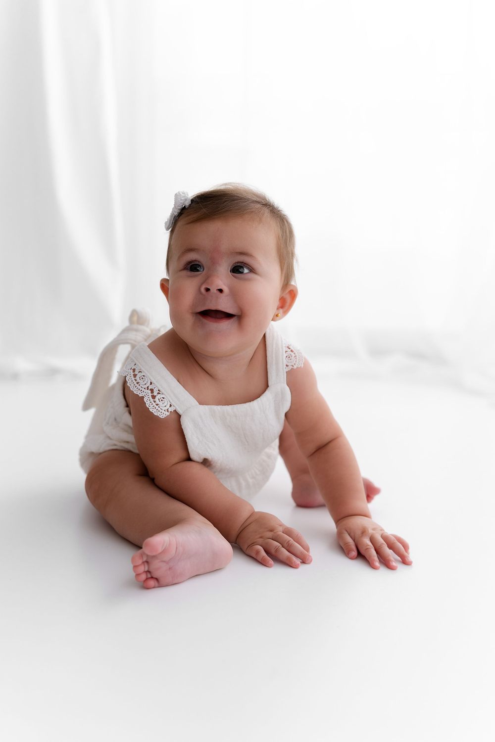 Smiling baby in white lace outfit sits during minimalist studio photography session.