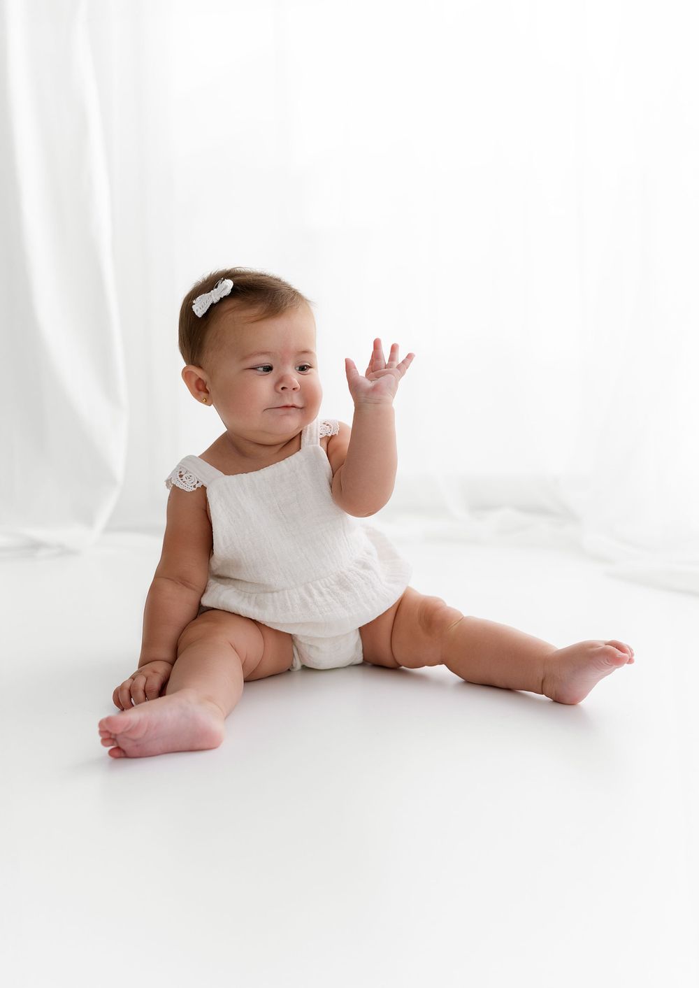 Baby in white outfit sits on white floor during minimalist studio photo session.