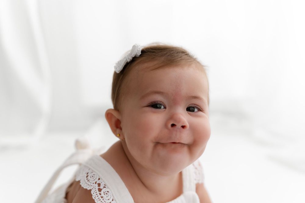 Baby in white lace outfit and bow headband smiling against white backdrop.