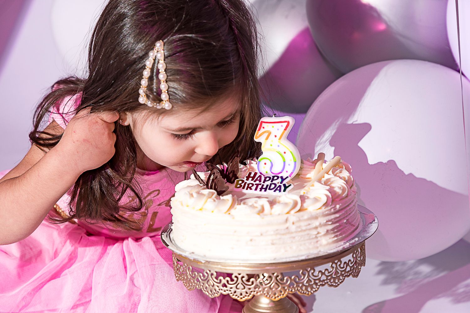 Baby girl in pink dress leaning over to taste birthday cake during photo session.