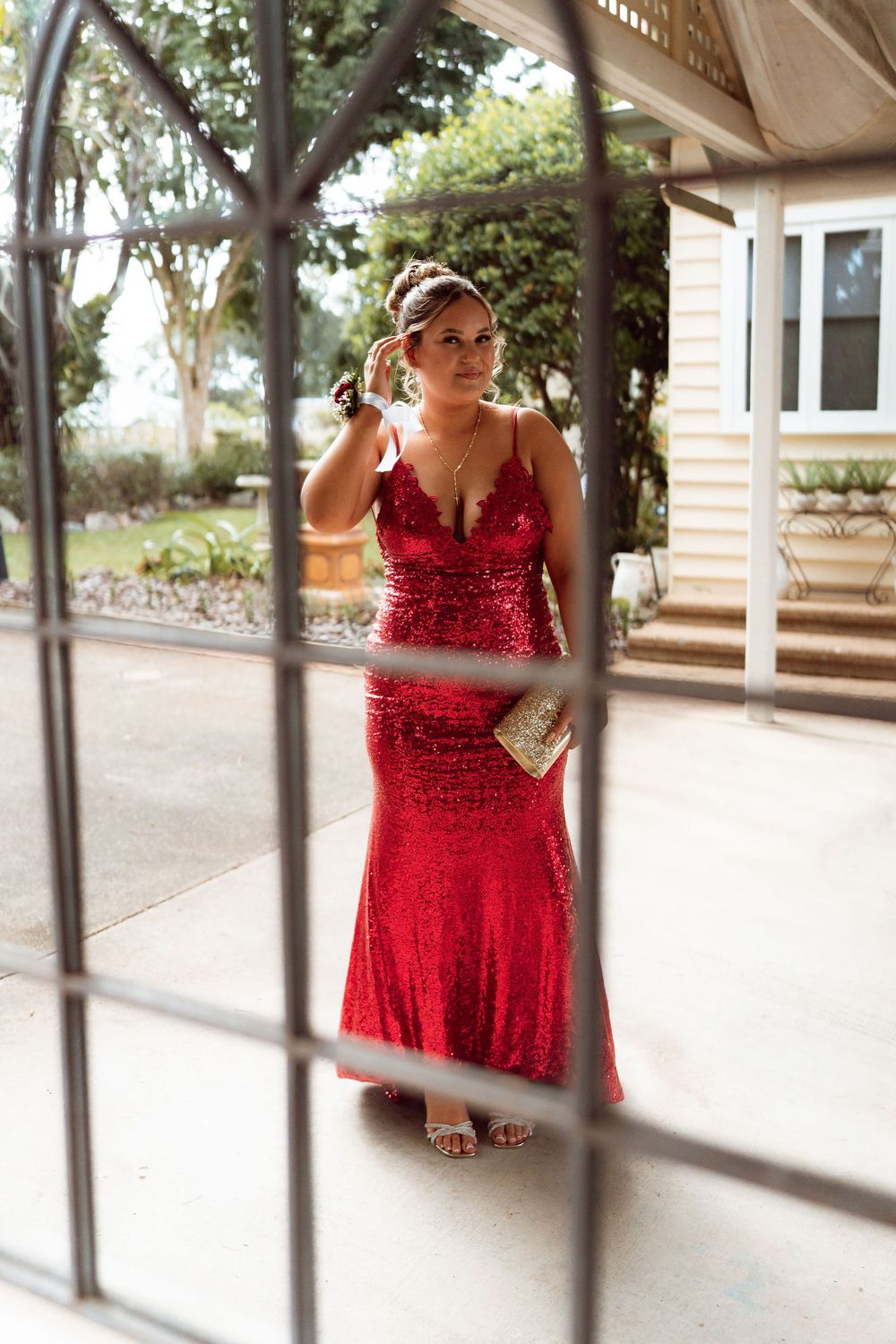 Woman in red sequin mermaid gown holding clutch, posing outdoors near a house for prom or formal event.