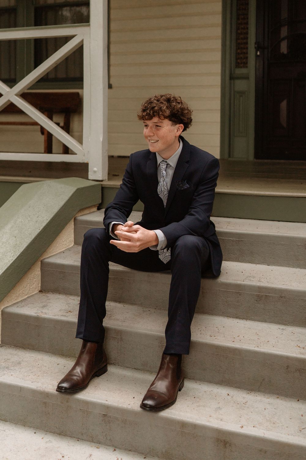 Young man in navy suit and brown boots sits smiling on concrete steps outside a classic building.