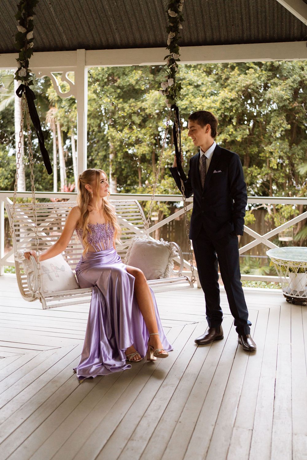 A young couple in formal attire on a porch, girl in lavender gown on swing, boy in dark suit standing nearby.