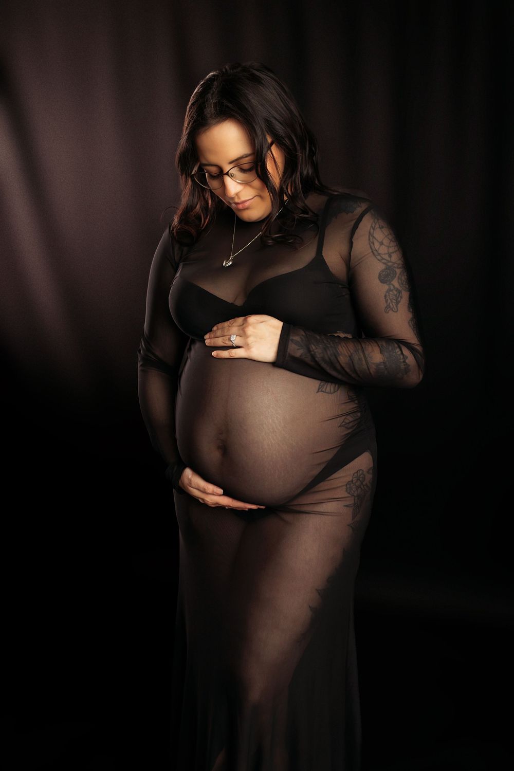 Pregnant woman in sheer black lace dress cradling baby bump in dramatic studio lighting.
