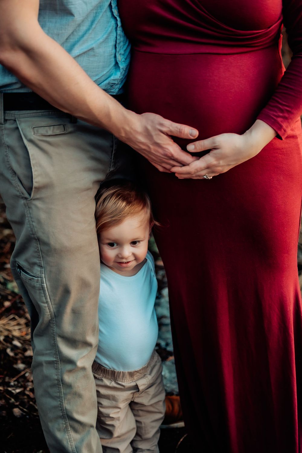 Close-up of expectant parents and toddler showing hands on pregnant belly in burgundy dress.