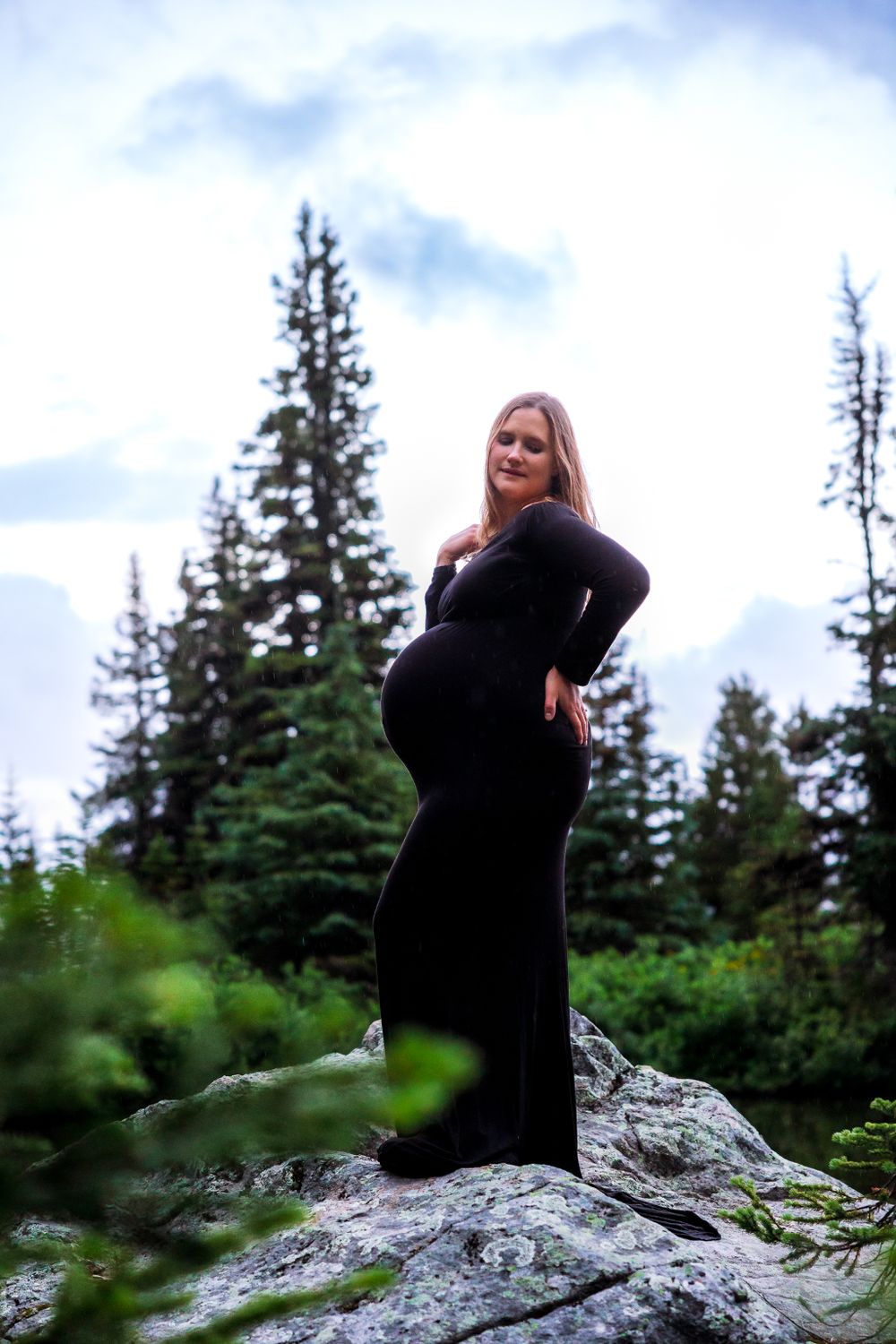Silhouette of pregnant woman in black dress standing on rocky outcrop surrounded by pine trees.