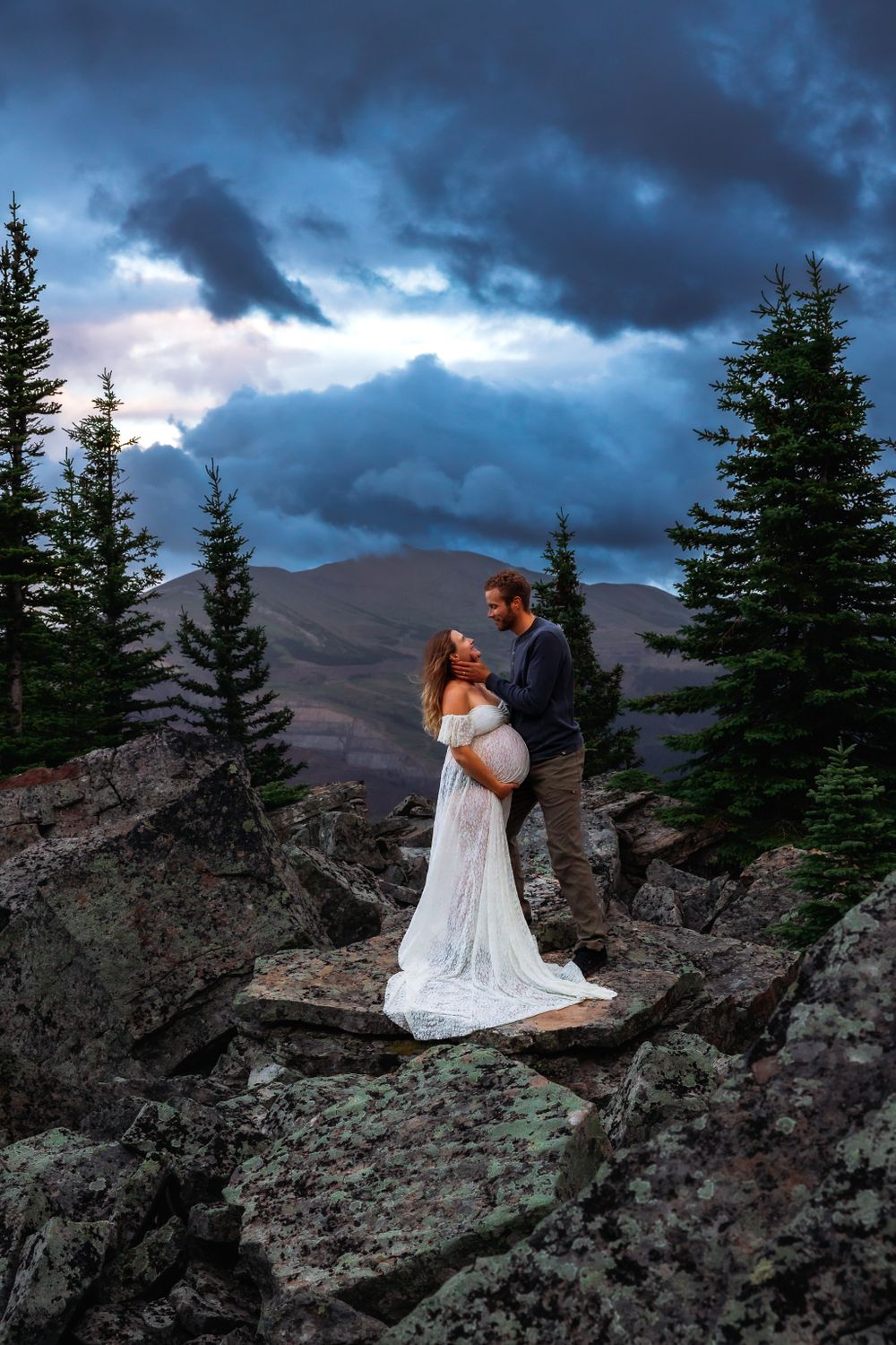 Dramatic mountain maternity photo with couple embracing against stormy sky and distant peaks.