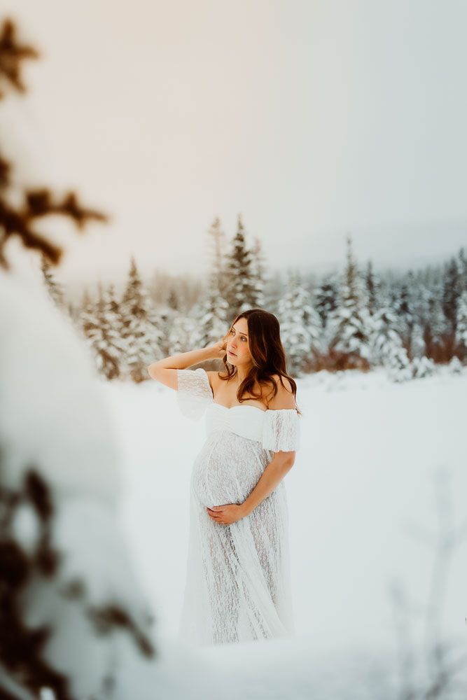 Maternity portrait in snowy winter landscape with woman in white lace dress.