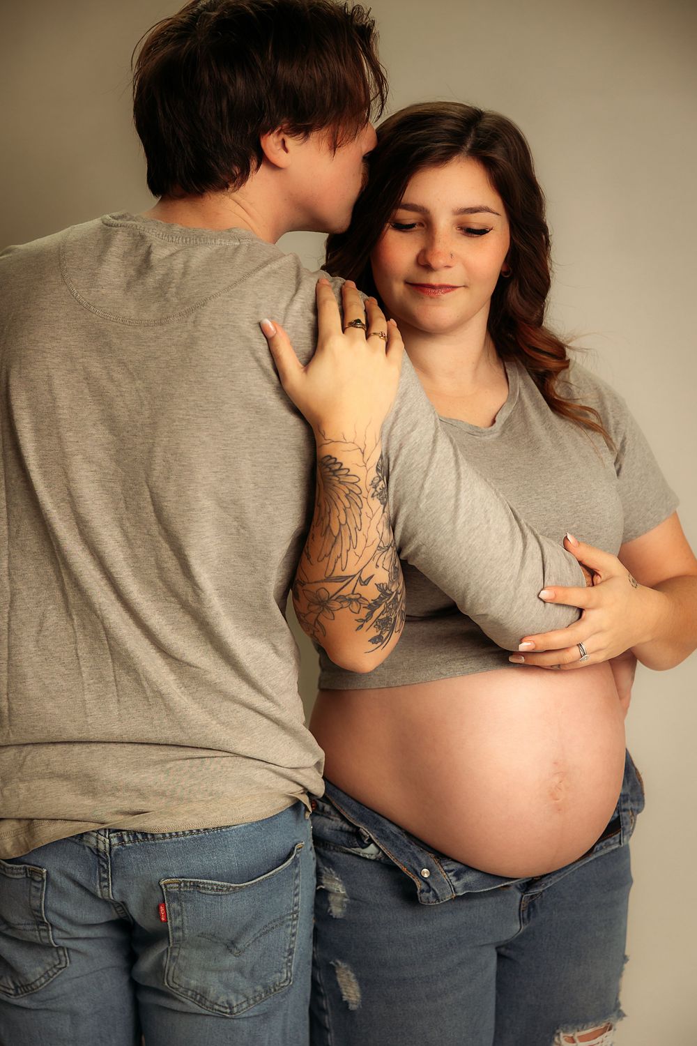 A tender maternity moment captured in casual wear as a couple embraces, showing a pregnant belly and visible tattoo.