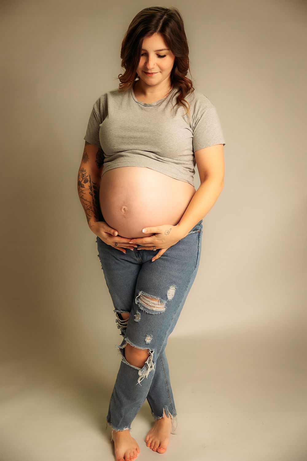 A casual maternity photo shoot showing someone in ripped jeans and a grey t-shirt against a neutral backdrop.