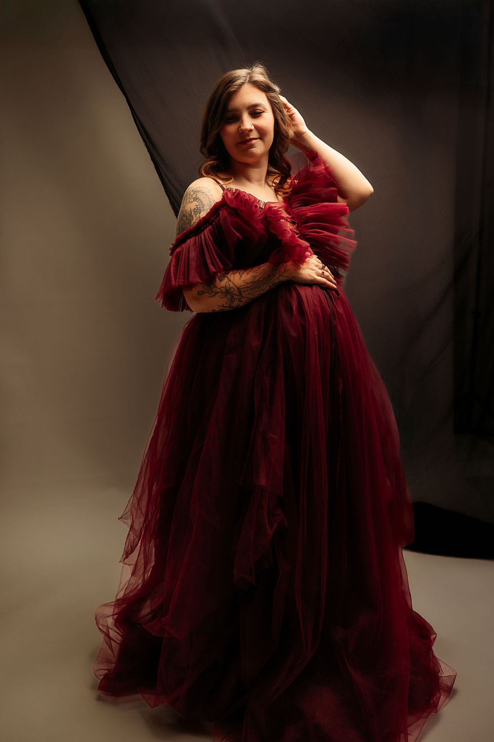 An expectant mother poses in a flowing burgundy tulle gown against a dark backdrop.