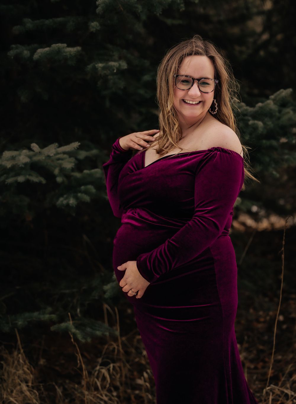 Moody maternity photos in a velvet burgundy dress against evergreen trees.