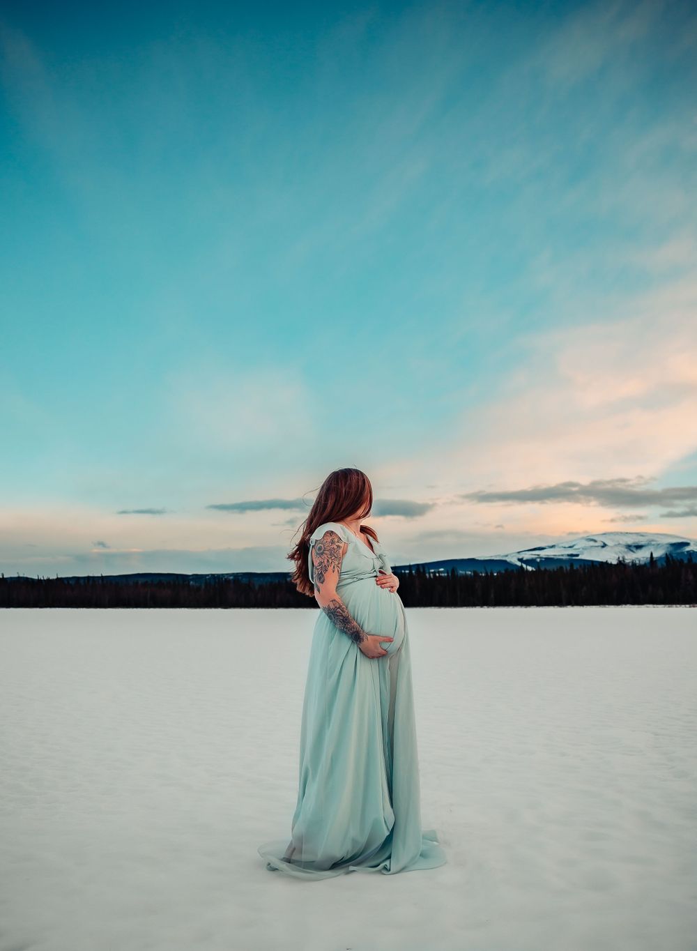 A person in a flowing mint dress stands against a snowy mountain landscape at sunset.