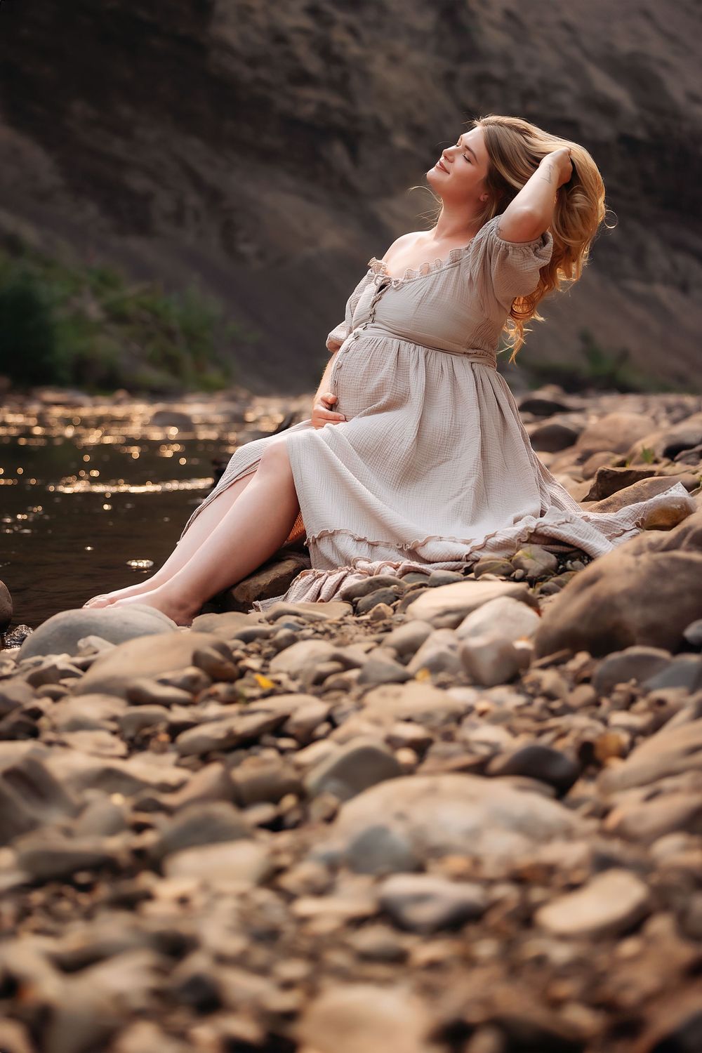 Artistic maternity photos on a rocky riverbank showing a woman in a flowing white dress at sunset.