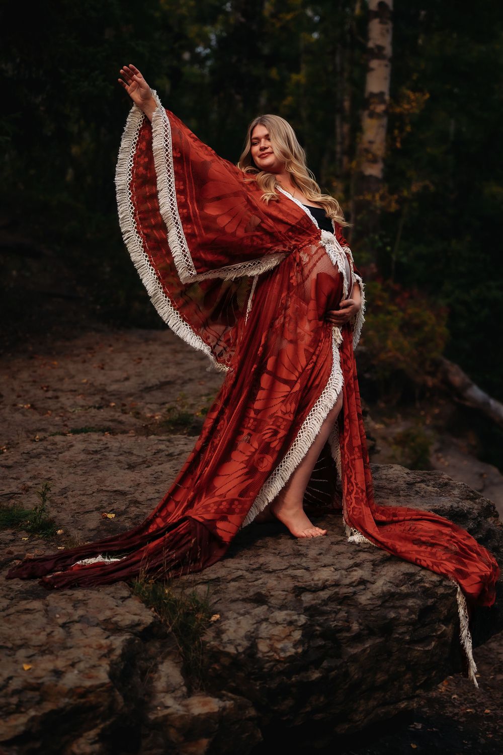Dramatic maternity photoshoot featuring a flowing red velvet dress with lace trim against a dark forest backdrop.