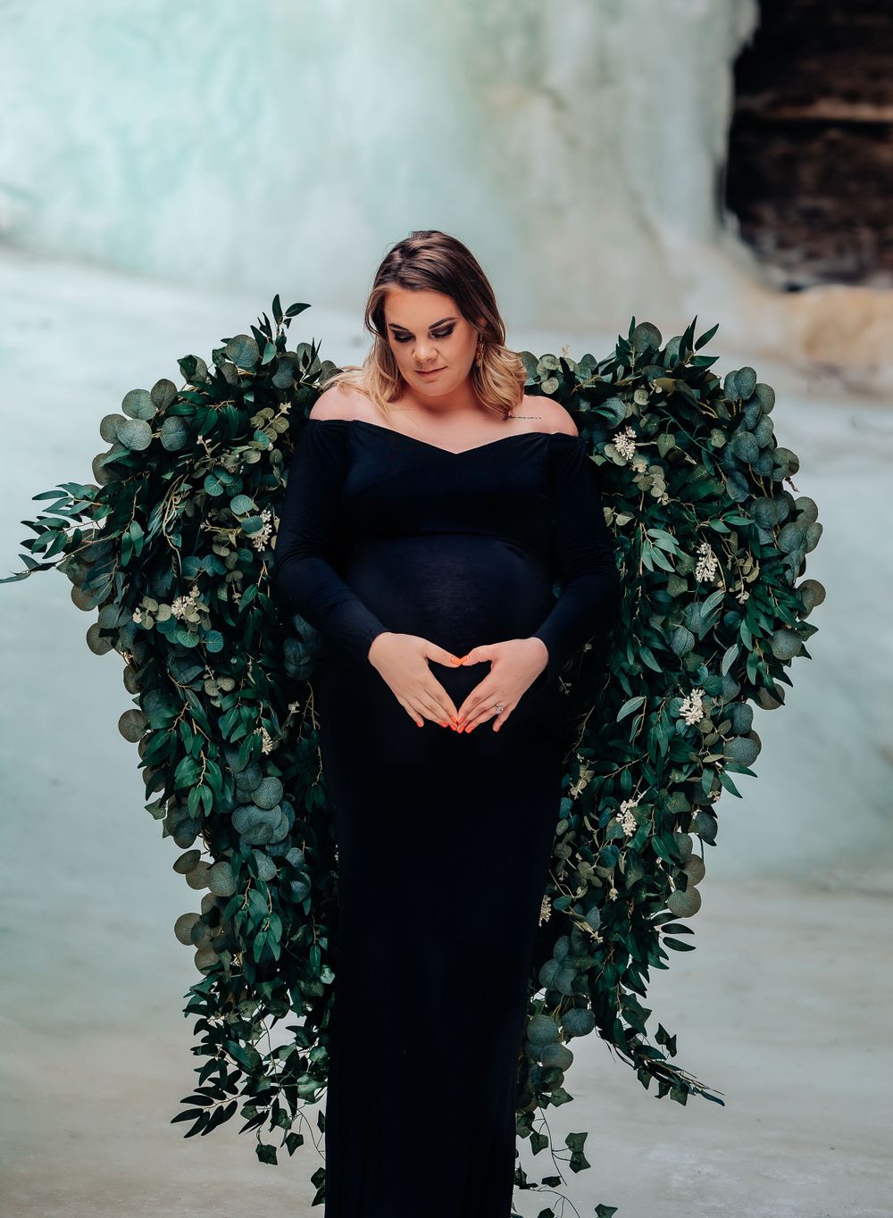Maternity portrait with leafy green wings and black gown against icy background.