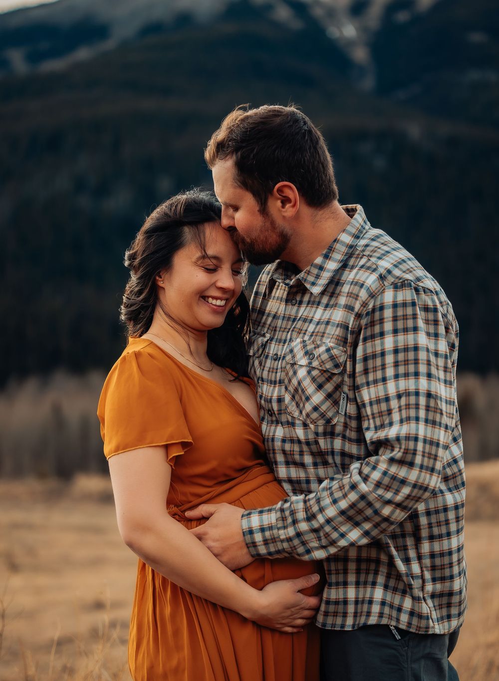 A couple in a field at sunset sharing an intimate moment as she wears a burnt orange dress and he wears a plaid shirt.